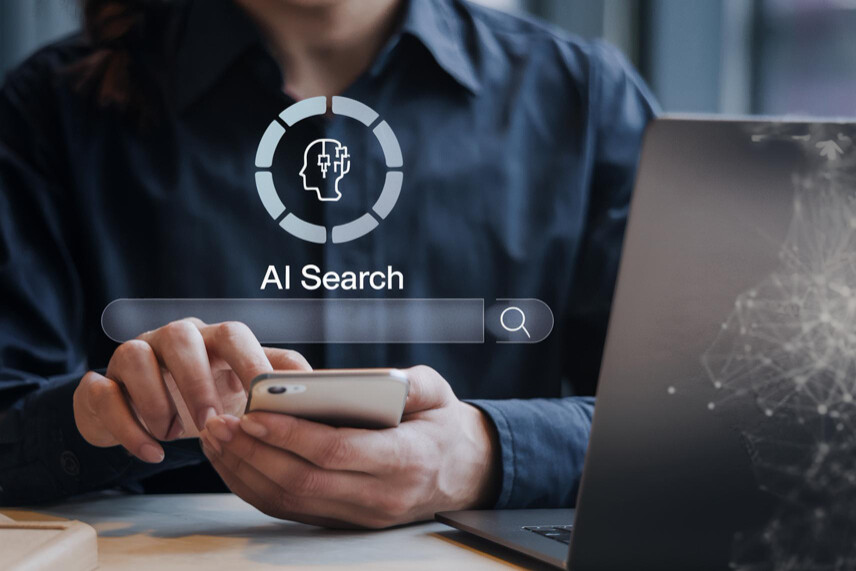 Άτομο χρησιμοποιεί smartphone δίπλα σε laptop με ένδειξη AI Search